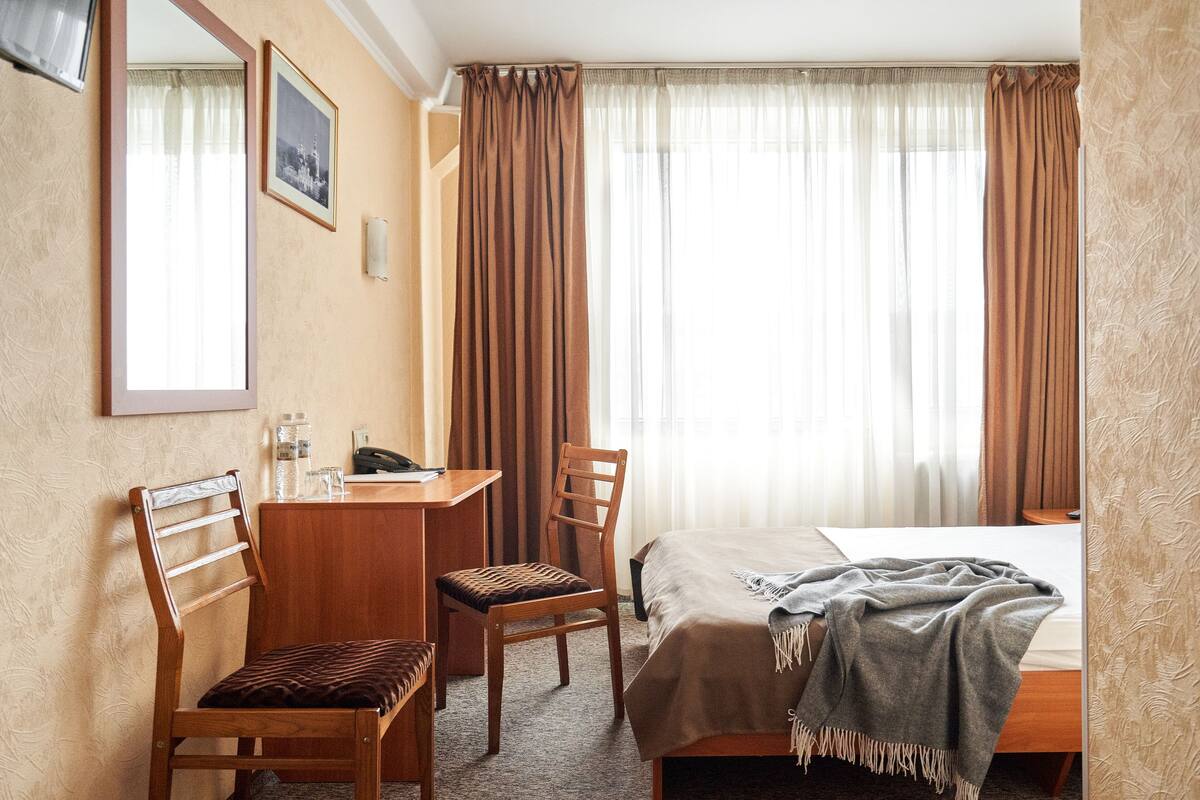 Ein Hotelzimmer mit Bett, Schreibtisch, zwei Stühlen, einem Spiegel, einem Bild an der Wand und einem Fenster mit Vorhängen.