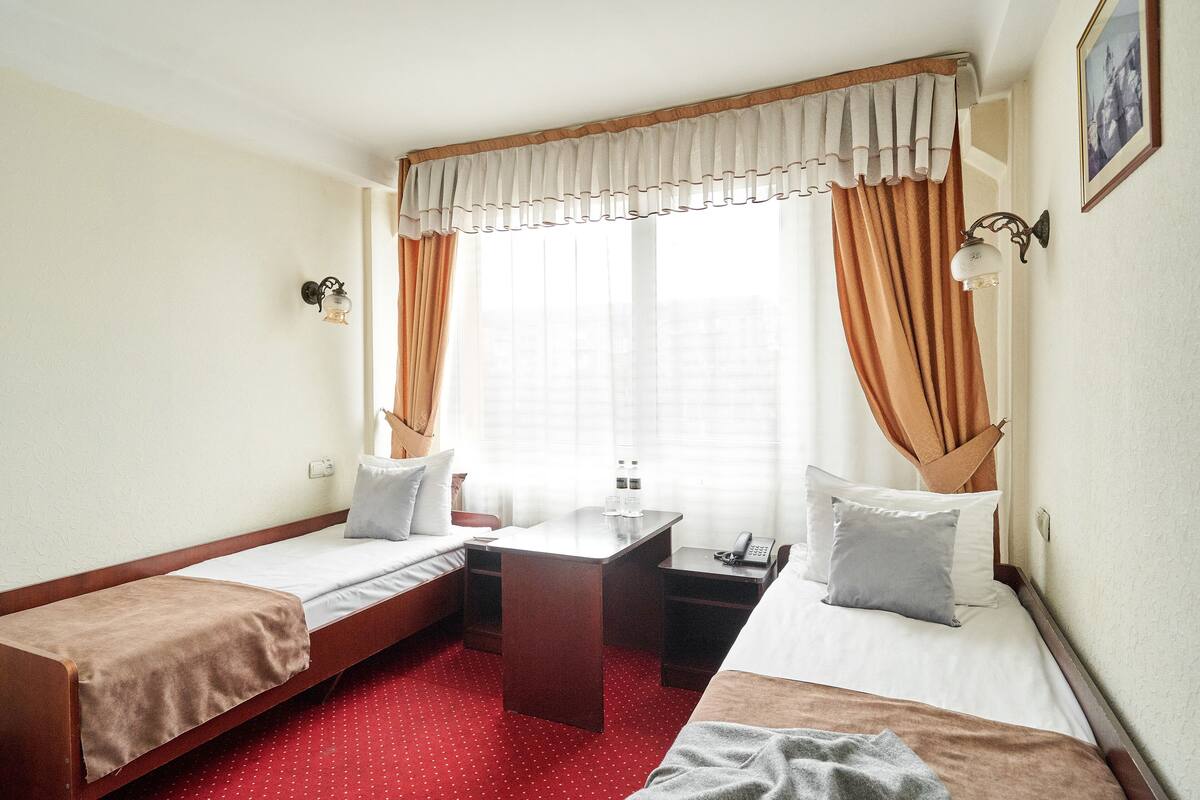 Ein Hotelzimmer mit zwei Einzelbetten, einem kleinen Schreibtisch, einem Telefon und einem fensterbepflanzten Fenster.