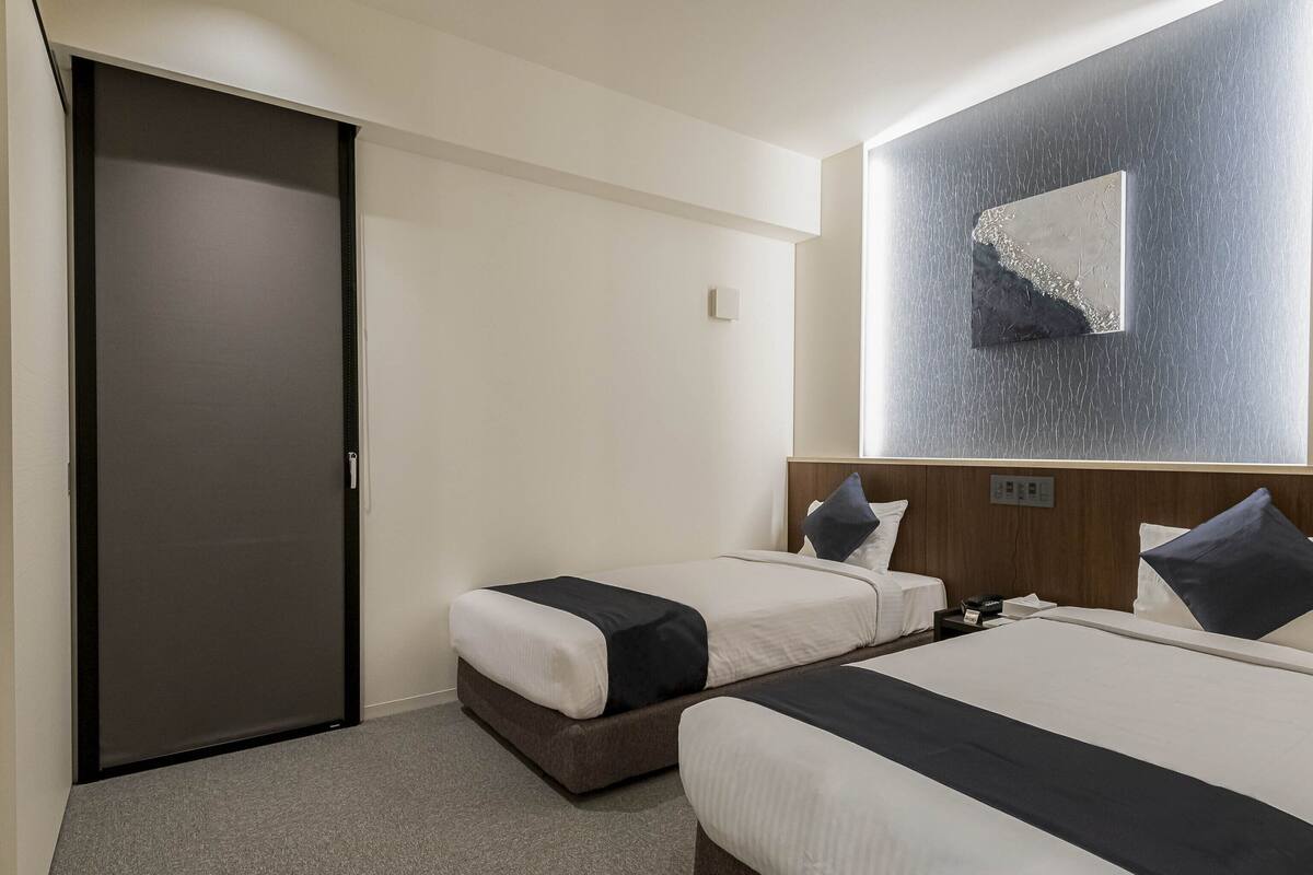 Une chambre d’hôtel avec deux lits, une porte sombre, un mur de couleur claire et un élément décoratif texturé sur le mur.