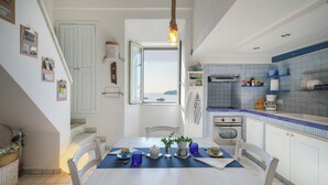 Un coin repas avec une table dressée pour deux personnes, une cuisine avec un plan de travail bleu et blanc à motifs, et une vue sur la mer par une fenêtre.