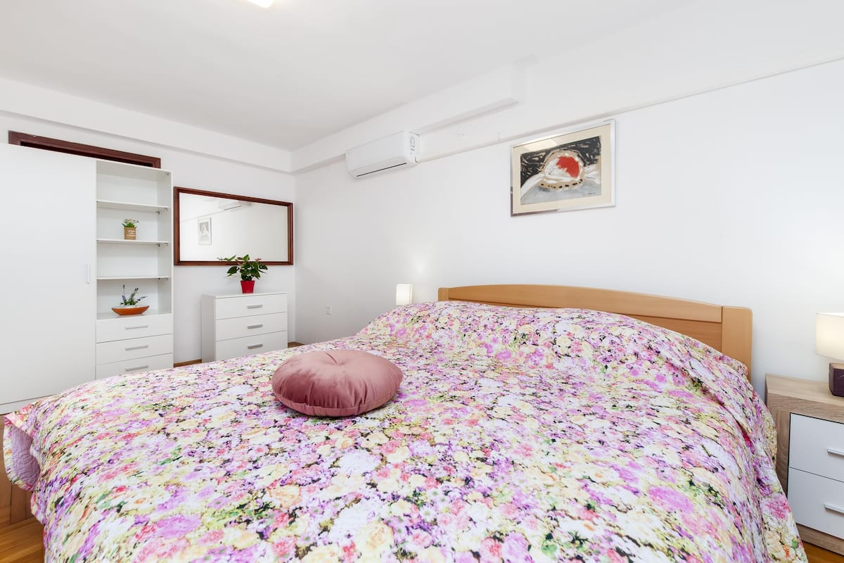 A bedroom with a floral bedspread, a wooden headboard, a bedside table with a lamp, a nightstand, a dresser, a mirror, and a potted plant.