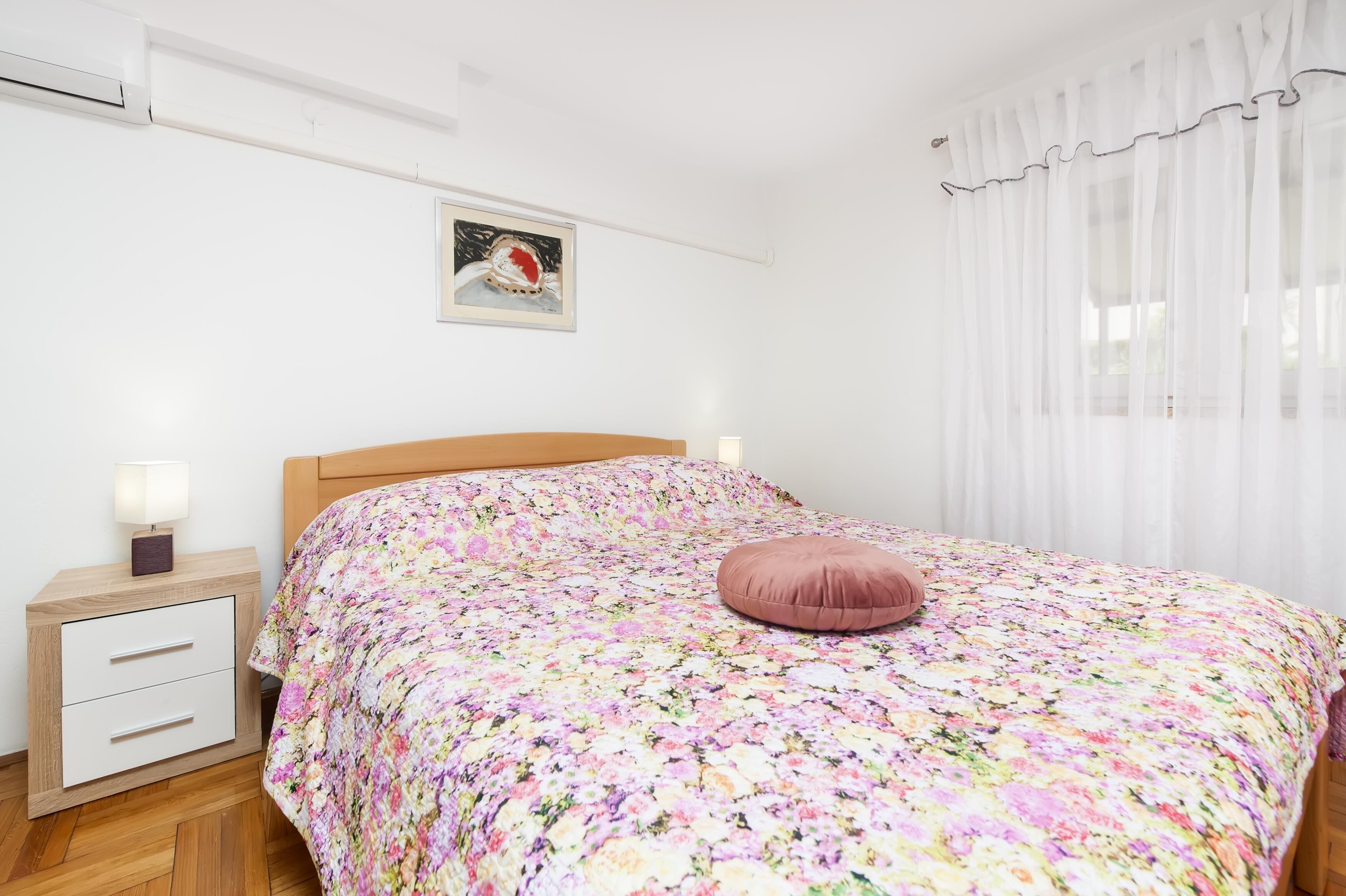 Um quarto com cabeceira de madeira, colcha florida, travesseiro rosa, mesa de cabeceira com abajur e quadro na parede.