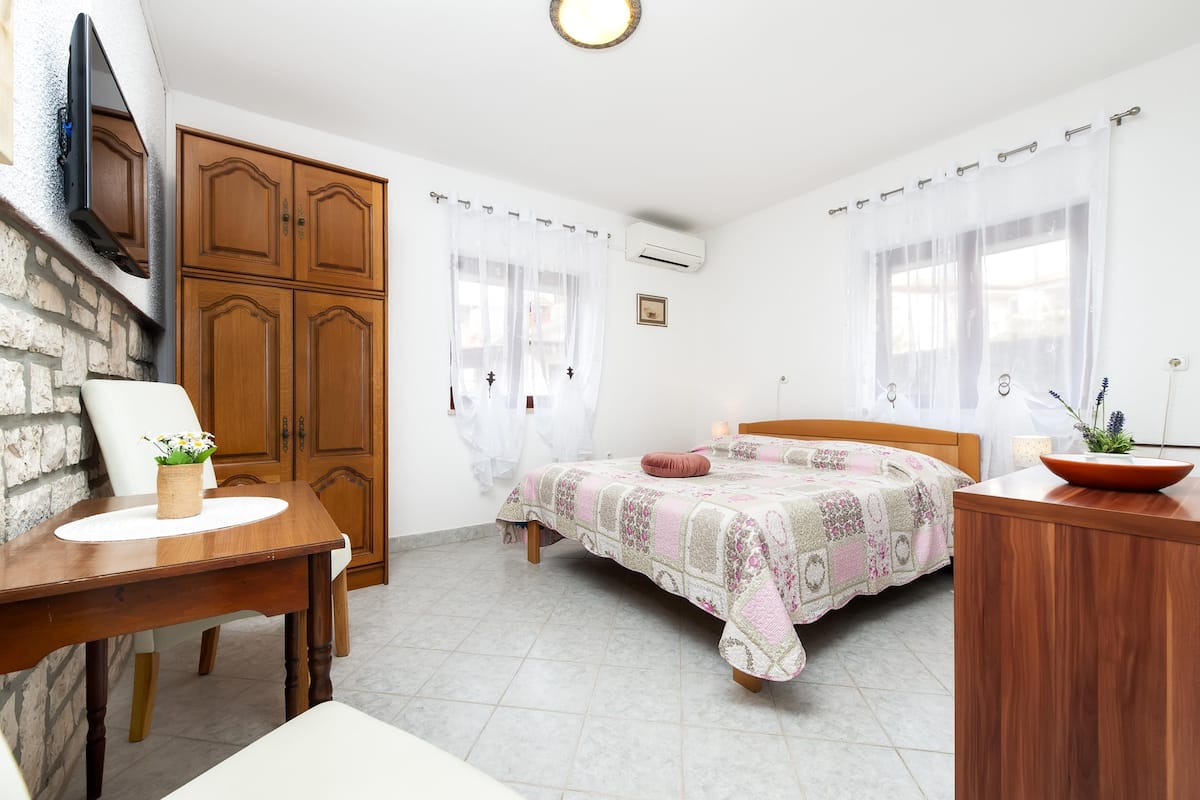 A bedroom with a wooden bed, a desk, and a chair. There is a stone wall and a wooden wardrobe.