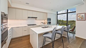Una cocina moderna con armarios blancos, una isla central con taburetes y un amplio ventanal con vistas a zonas verdes.
