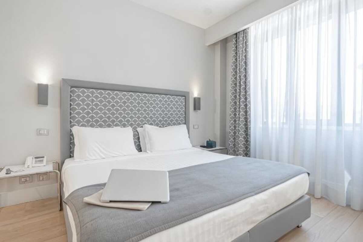 A modern hotel room with a large bed, a laptop on the bed, a bedside table, and a nightstand.