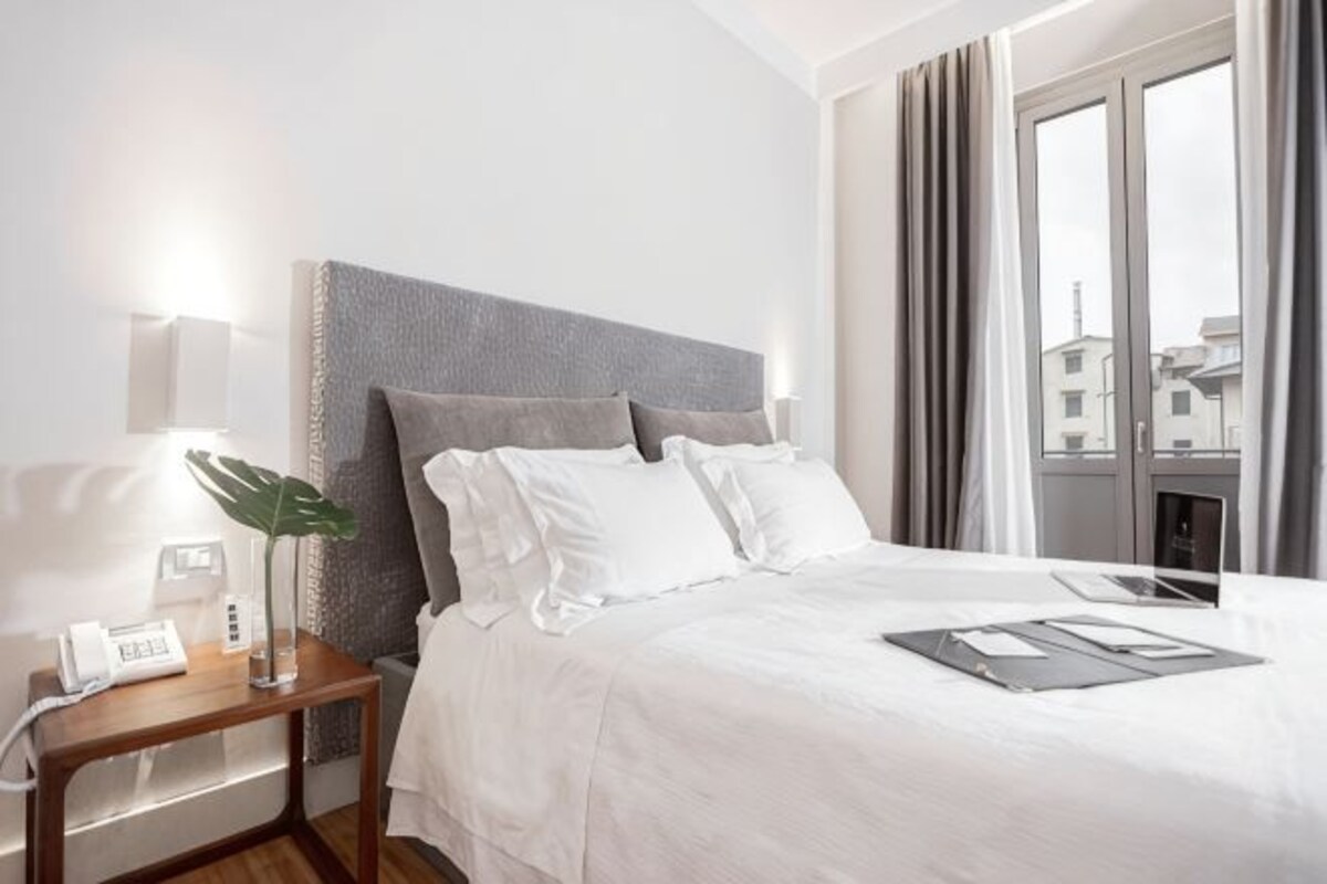 A hotel room with a large bed, a bedside table with a lamp, a phone, and a window with curtains.