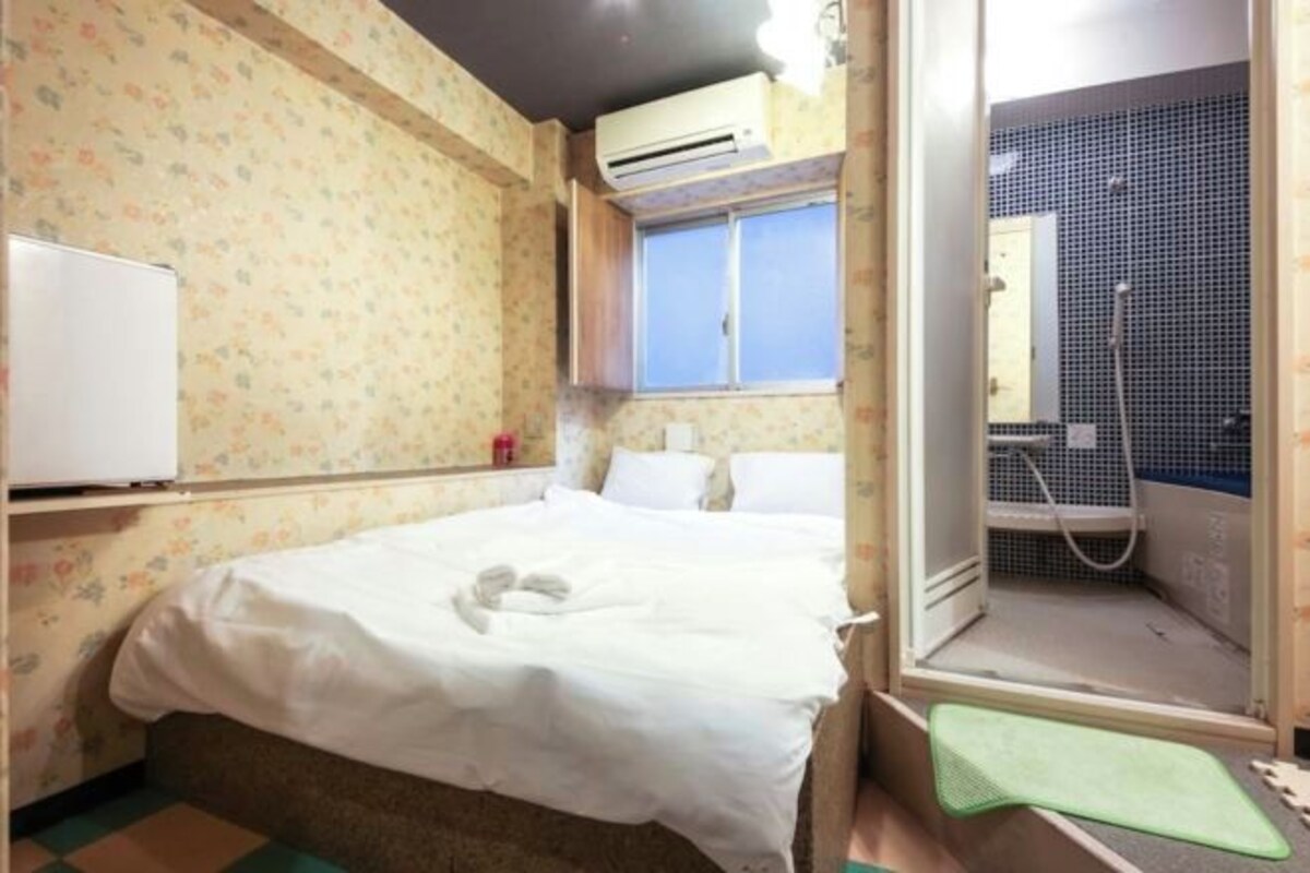 Quarto pequeno com uma cama de solteiro, parede com padrão floral, janela com vista e área de chuveiro.