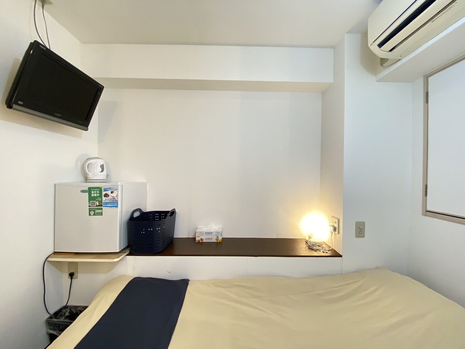 Une chambre d’hôtel de taille réduite, comprenant un lit, un petit bureau avec une lampe, une télévision, un réfrigérateur et un appareil de climatisation.
