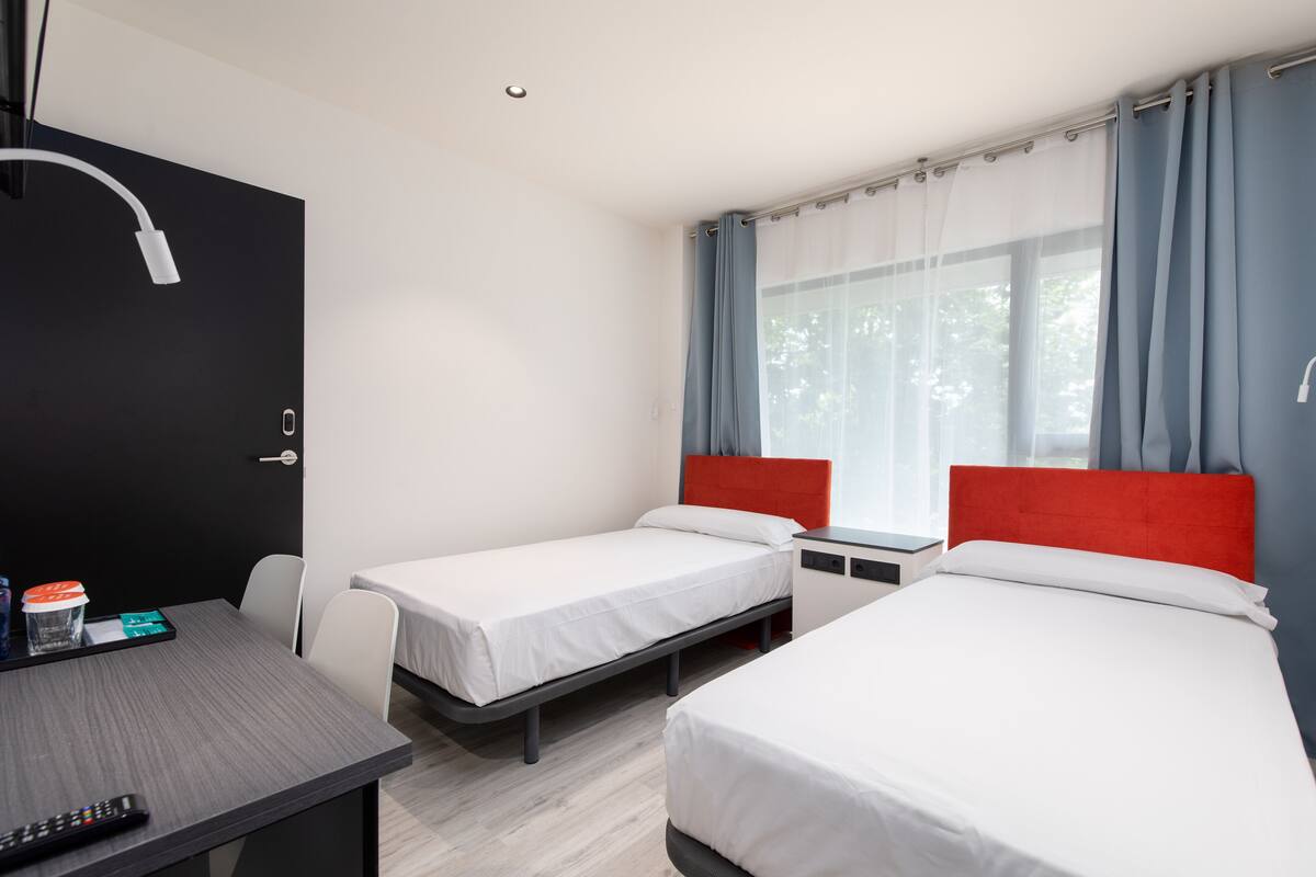 Habitación de hotel con dos camas individuales, un escritorio con silla, una mesita con control remoto y una ventana con cortinas.