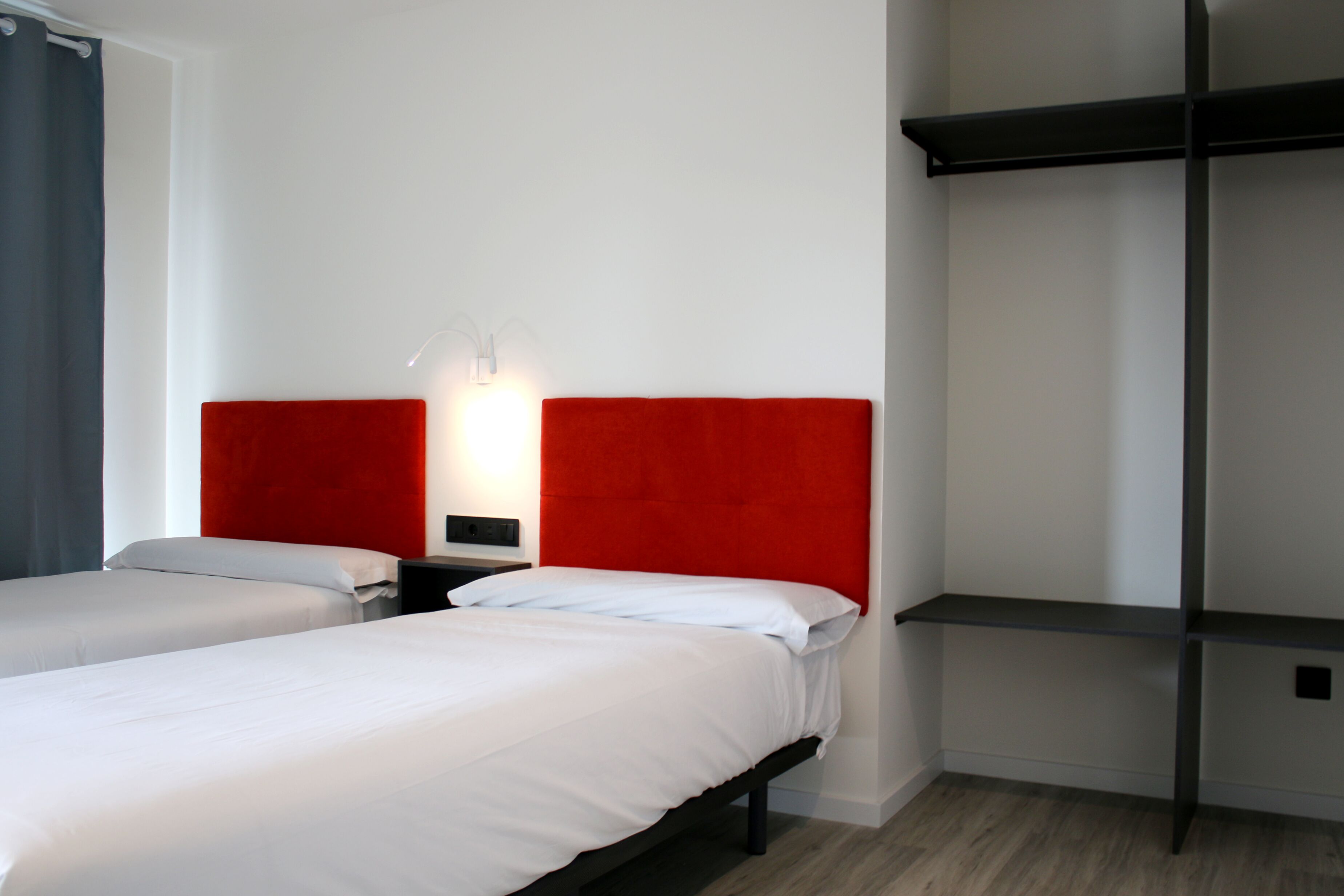 Habitación de hotel con dos camas, cabecero rojo, mesita de noche y estante de pared.