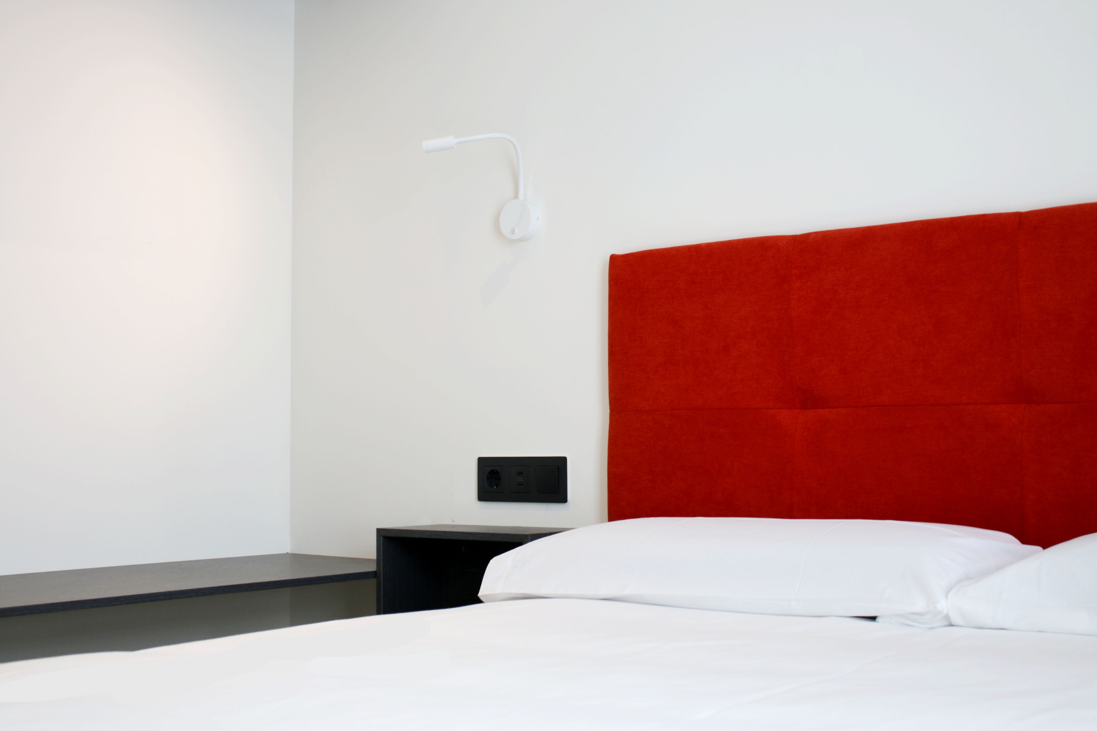 A modern hotel room with a red headboard, white bedding, a bedside table, and a wall-mounted light fixture.