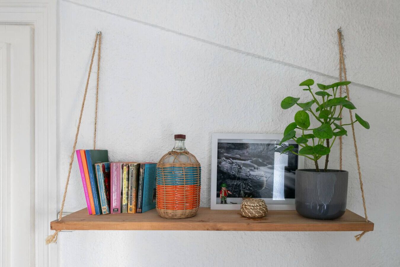 Une étagère en bois, suspendue à une corde, sur laquelle sont posés des livres, une bouteille, un tableau encadré et une plante en pot.