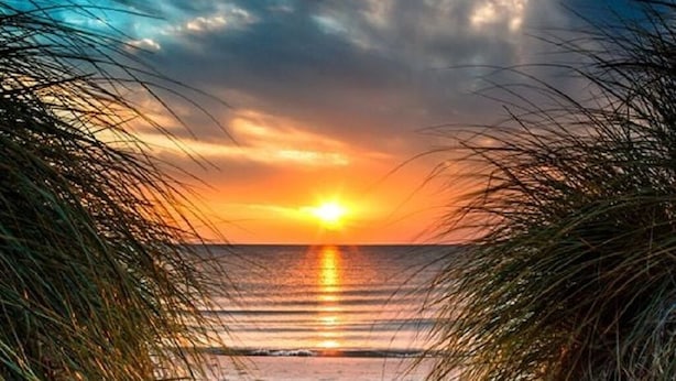 Un sentier qui longe la plage et mène à l’océan, avec un coucher de soleil en arrière-plan.