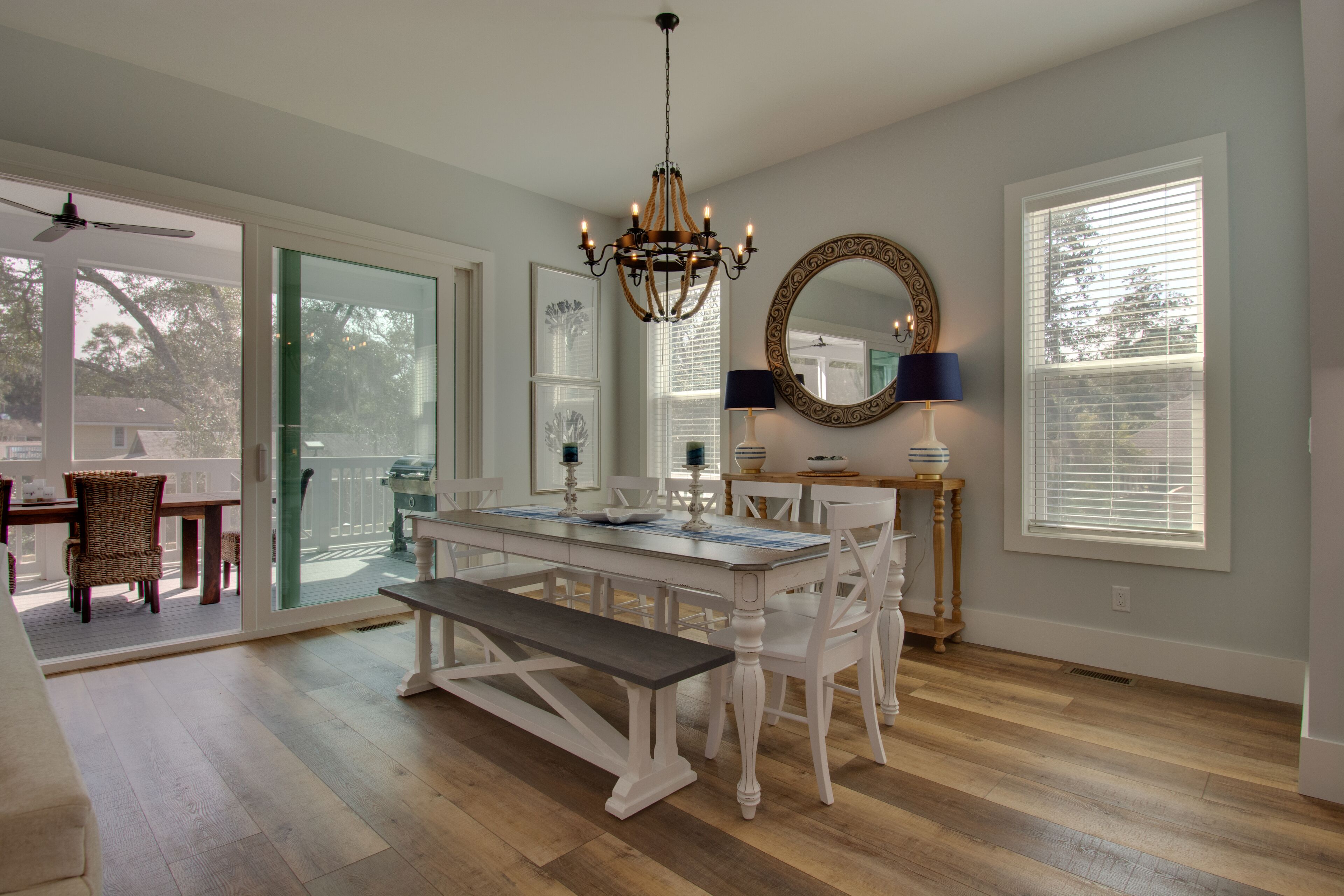 A dining area with a white table, bench, and chairs, a chandelier, a mirror, and a window with blinds.