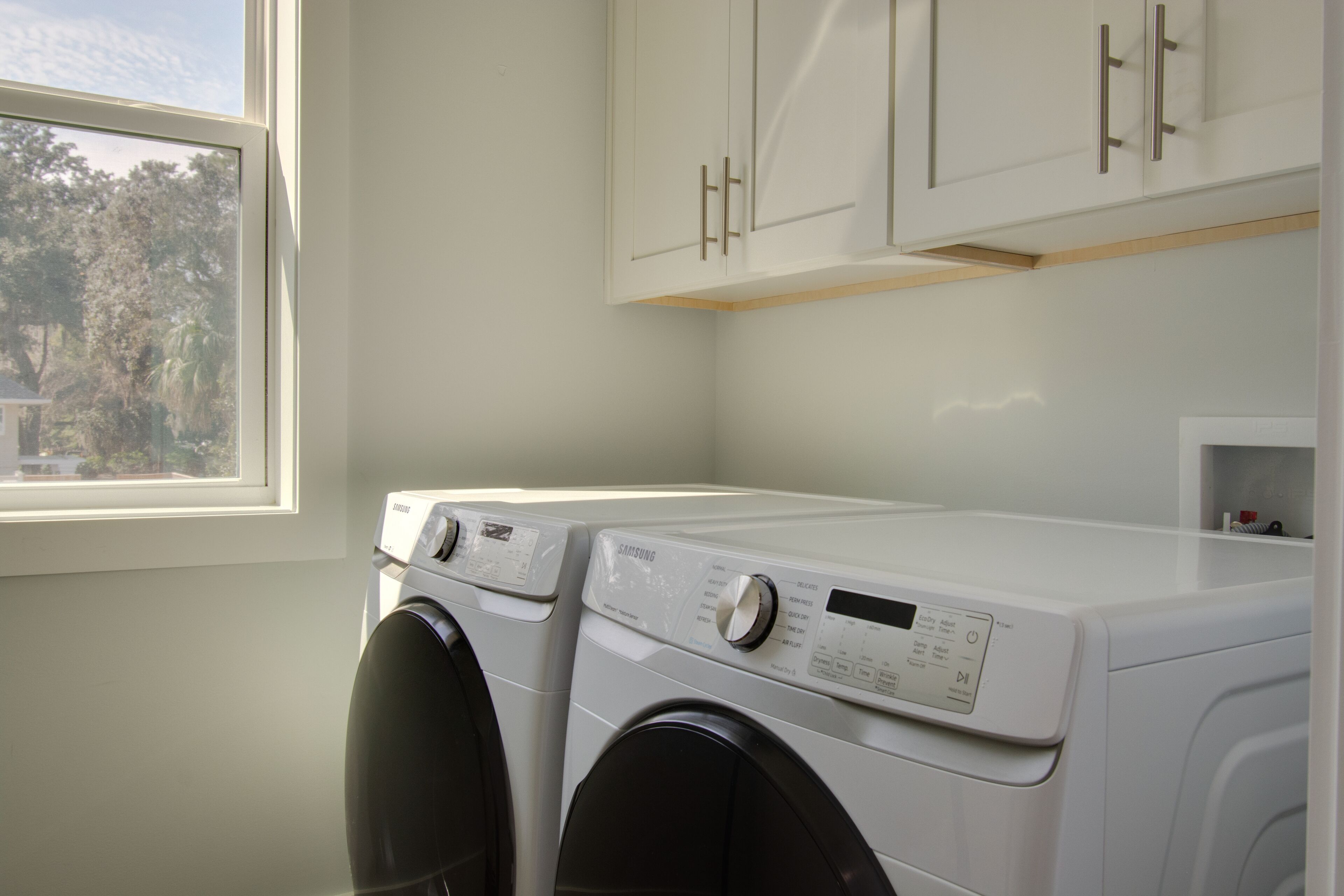 A laundry room with two front-loading washing machines, wooden cabinets above, and a window providing natural light.