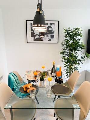 A dining area with a glass table, two chairs, a black pendant light, a framed artwork, a potted plant, a bottle of champagne, glasses, a newspaper, and a croissant. - Tudors eSuites Luxury House Garden (Birmingham)