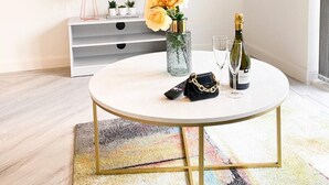 A modern living room with a flat-screen TV mounted on the wall displaying 'NETFLIX'. There is a white round coffee table with a decorative vase, a bottle of champagne, and a handbag. The room has a minimalist design with a grey console table and a colorful rug. - Tudors eSuites Luxury House Garden (Birmingham)
