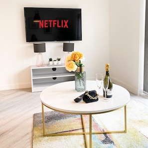 Ein modernes Wohnzimmer mit einem flachbildfernseher an der Wand, der 'NETFLIX' anzeigt. Ein weißer runder Couchtisch ist mit einer dekorativen Vase, einer Champagnerflasche und einer Handtasche bestückt. Das Zimmer hat ein minimalistisches Design mit einem grauen Konsolentisch und einem farbenfrohen Teppich.