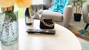 A living room with a round table, a vase of flowers, a remote control, and a sofa with a blue throw blanket. - Tudors eSuites Luxury House Garden (Birmingham)