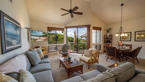 A spacious living room with a large sofa, a coffee table, and a dining area with chairs and a table. There is a ceiling fan, large windows, and artwork on the walls. - Private and Sweet Wailea 1 BD+2 BA Grand Champions Villa (Wailea)