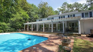 A pool with a brick deck, a covered patio with chairs, and a white building with columns. - 4 Bedroom Pinehurst Home with Pool and Hot Tub (Pinehurst)