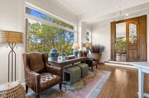 A spacious room with a large window, leather armchair, wooden table, and a view of trees outside.
