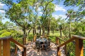 An outdoor deck with a round table and chairs, surrounded by a wooden railing, overlooking a forested area.
