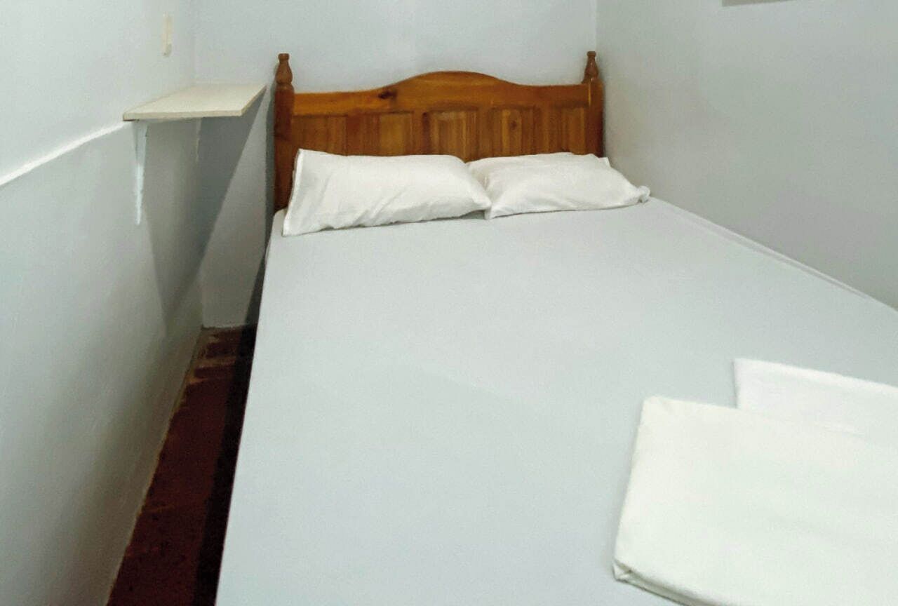 Een kleine kamer met een enkel bed, een houten hoofdbord, witte beddengoed en een klein nachtkastje.