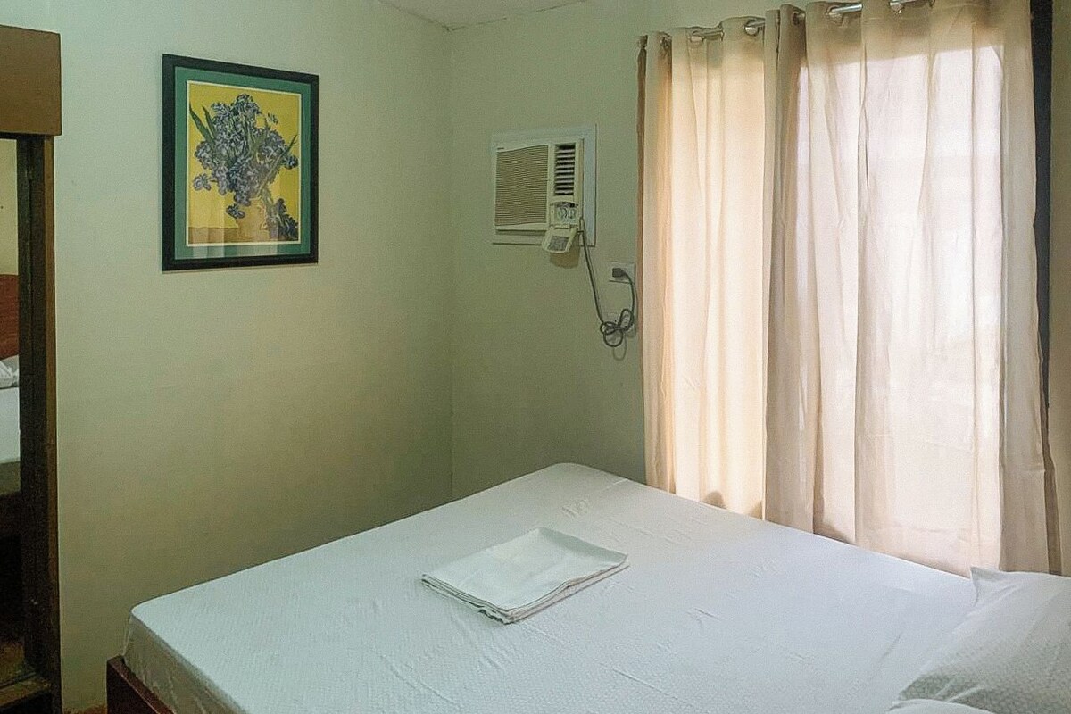 A single bed with a white sheet, a framed picture on the wall, a window with curtains, and a wall-mounted air conditioning unit.