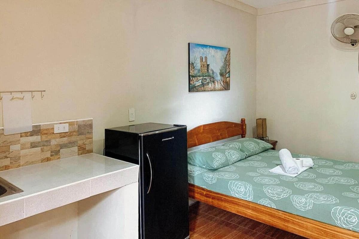 Une petite pièce propre, avec un lit, un réfrigérateur, un tableau au mur et un ventilateur.