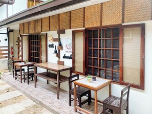 Un espacio al aire libre de estilo japonés tradicional, con mesas y sillas de madera, un pequeño jardín y un edificio con suelos de tatami y puertas shoji.