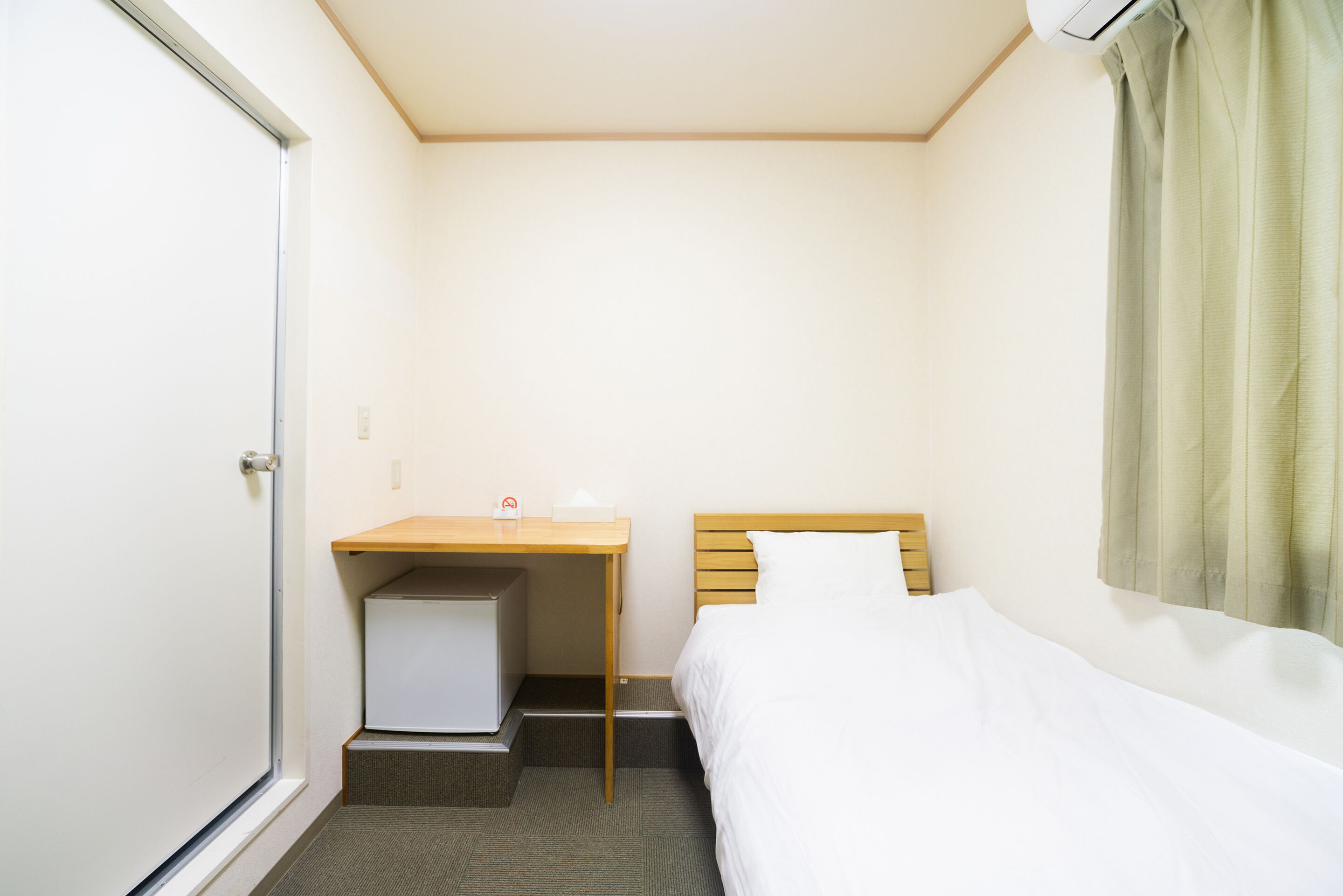 Une petite chambre d’hôtel propre, avec un lit simple, un bureau avec une chaise et un petit meuble de rangement sous le bureau.