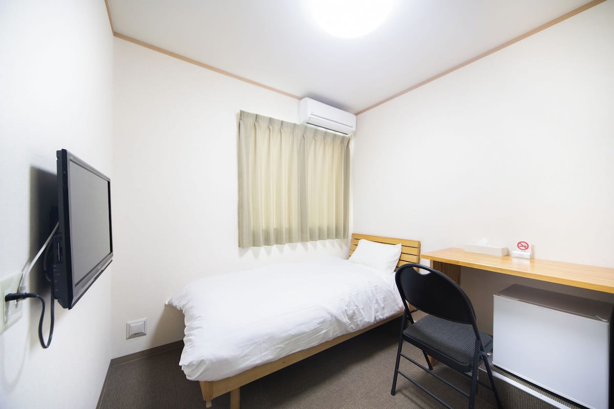 Ein kleines, sauberes Zimmer mit einem Bett, einem Schreibtisch, einem Sessel und einem Fernseher.