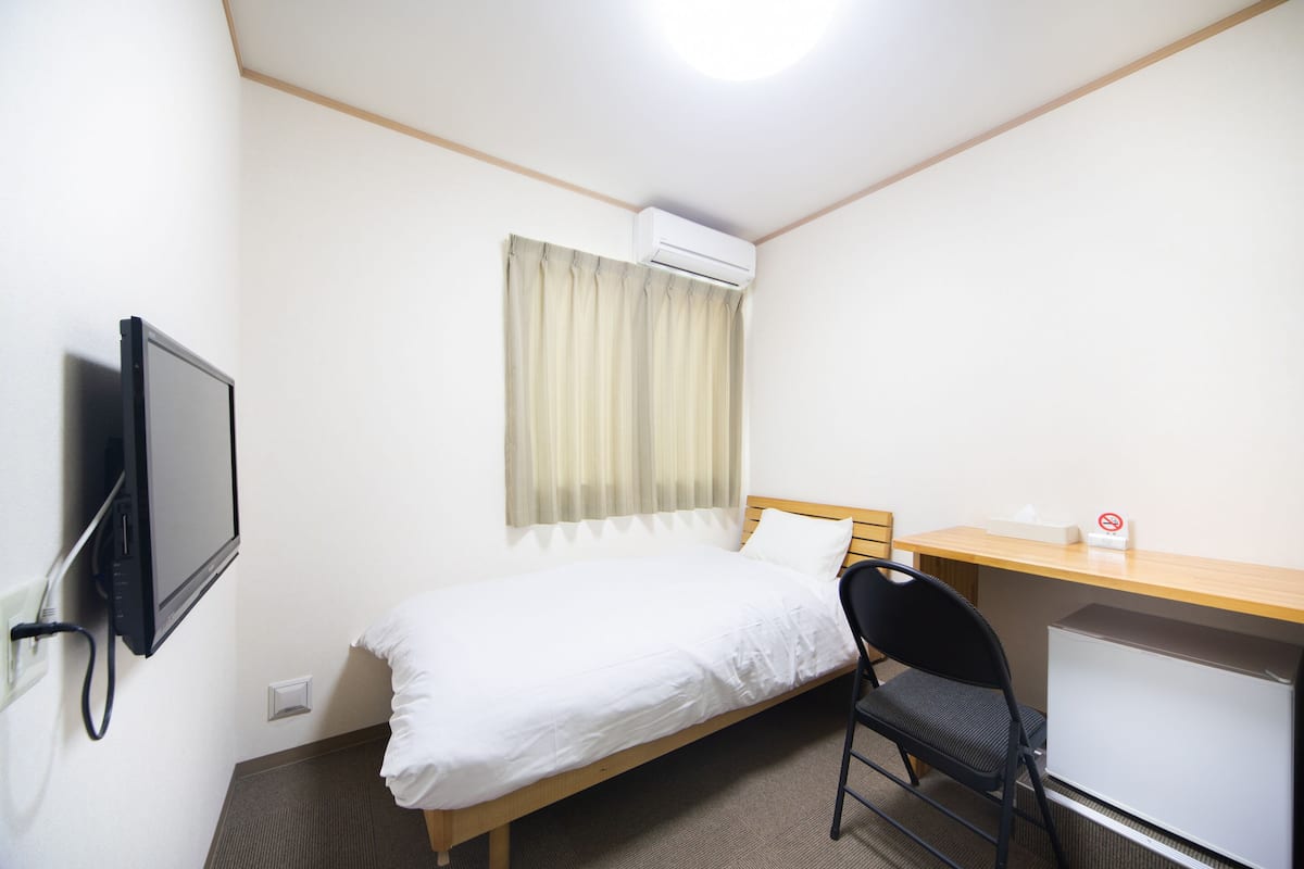 Ein kleines, sauberes Zimmer mit einem Bett, einem Schreibtisch, einem Sessel und einem Fernseher.