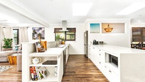A modern kitchen with white cabinetry, a wooden floor, and a dining area with a view of the outdoors. - Seven Smiles, Gerroa, South Coast (Gerroa)