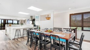 A modern kitchen with a central island, bar stools, and a dining area with a wooden table and chairs. - Seven Smiles, Gerroa, South Coast (Gerroa)