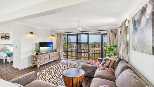 A spacious living room with a large TV, a sofa, a coffee table, and a view of the beach through large windows. - Seven Smiles, Gerroa, South Coast (Gerroa)