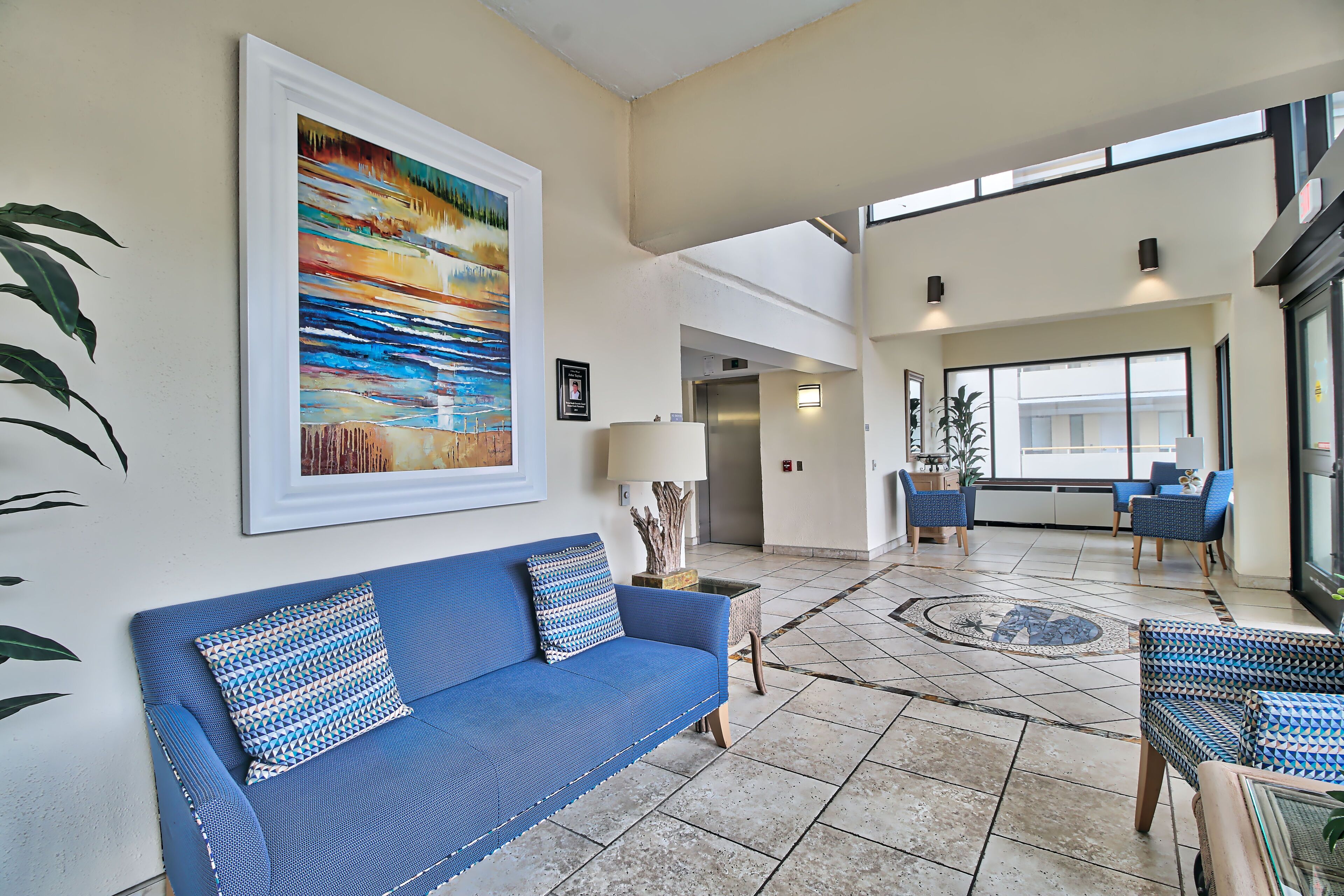 A spacious lobby with a blue sofa, patterned cushions, a large abstract painting, and a view of the outside through large windows.