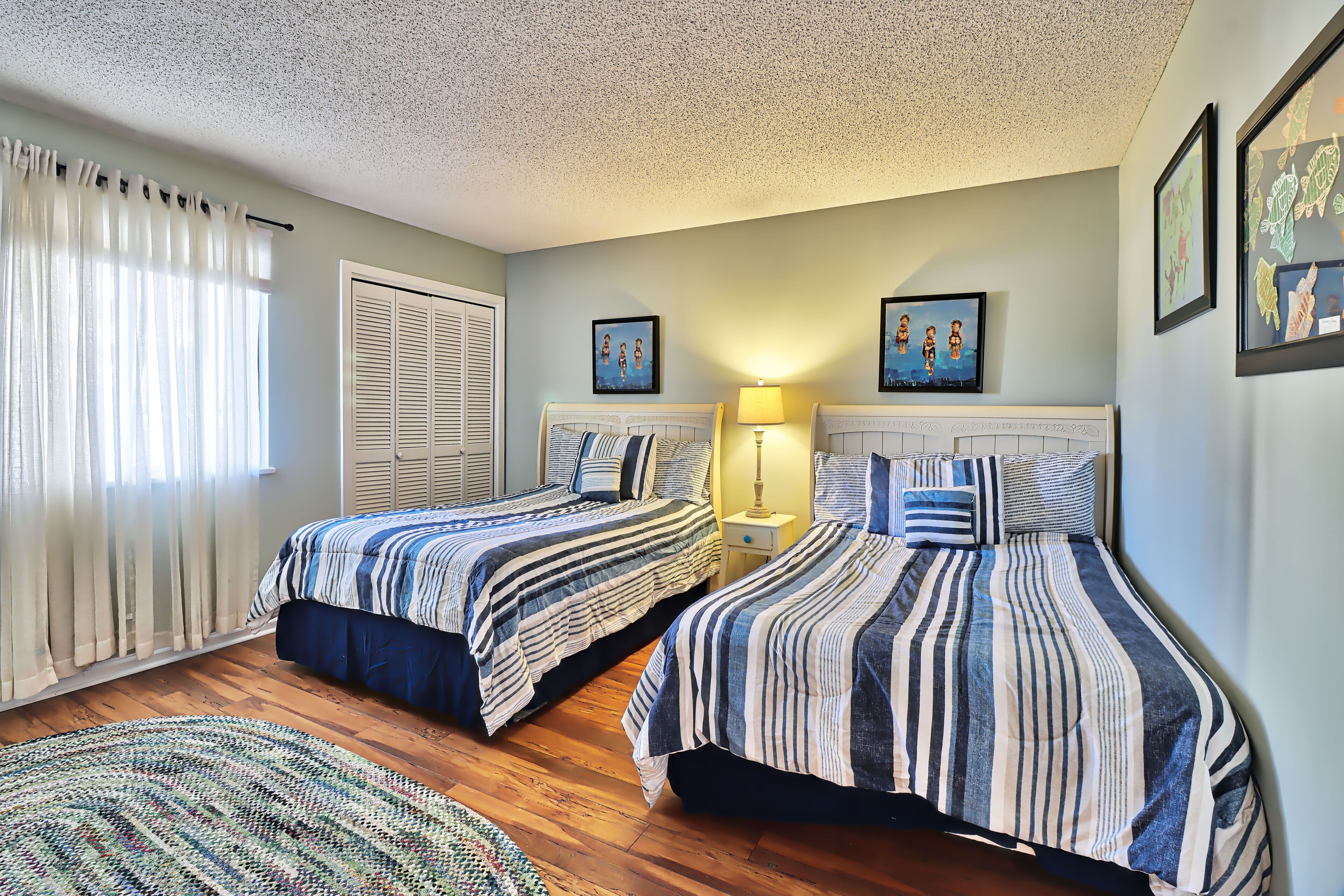 A bedroom with two beds, a window with curtains, a door, and a wall with framed pictures.
