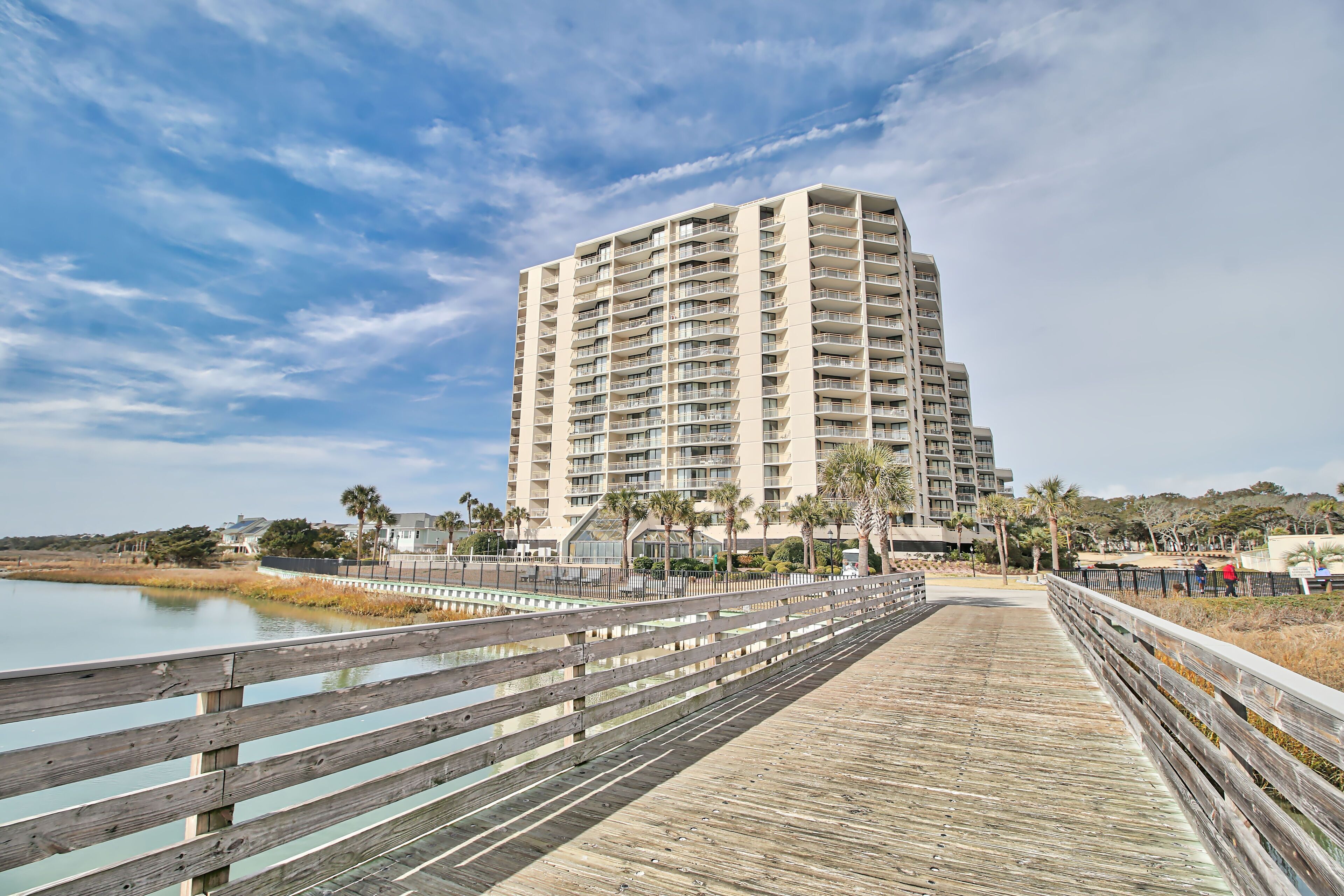 A tall, multi-story hotel with a wooden boardwalk leading to a body of water, surrounded by palm trees and a clear sky.