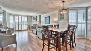 A spacious living area with a wooden dining table, comfortable seating, large windows, and a ceiling with a textured finish. -  Golfers Welcome, South Tower-Ocean Creek-1st Floor-Spacious Condo,Openings (Myrtle Beach)