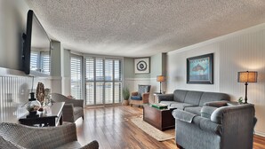 A spacious living room with a large gray sofa, a wicker chair, and a coffee table. There is a television mounted on the wall and a framed picture above the sofa. -  Golfers Welcome, South Tower-Ocean Creek-1st Floor-Spacious Condo,Openings (Myrtle Beach)