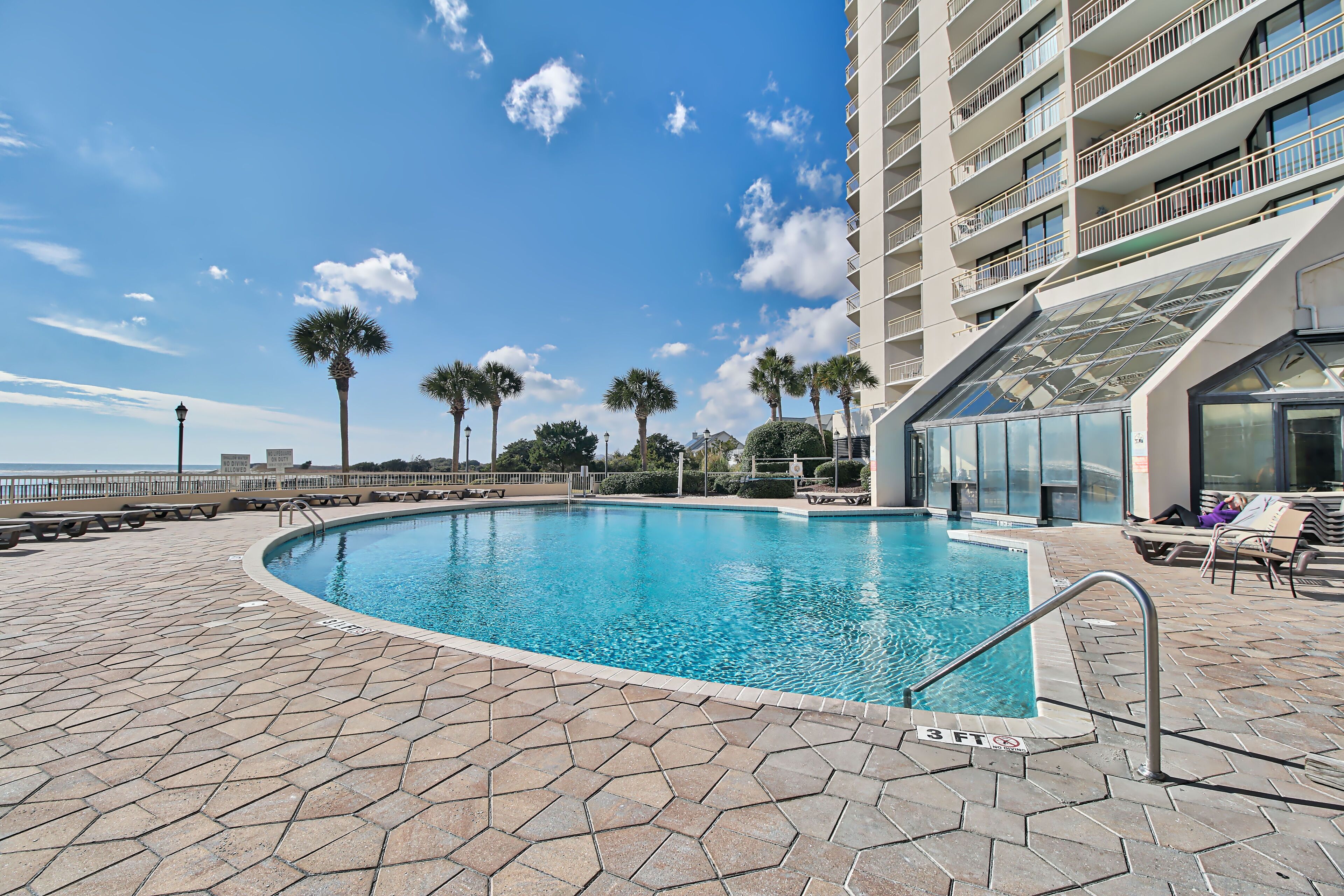 A swimming pool with a 3ft depth marker, surrounded by a tiled deck, palm trees, and a high-rise building with balconies.