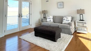 A spacious living room with a grey sofa, a black ottoman, and a white rug. There are two bedside tables with lamps and a framed picture on the wall. - Luxury Sea Bright Home, Ocean Views, 6 Sun Decks! (Sea Bright)
