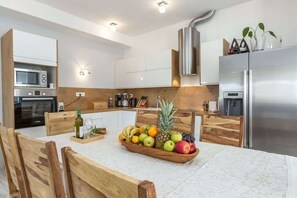 A modern kitchen with wooden cabinets, a stainless steel refrigerator, and a fruit bowl on the dining table. - Beachfront Holiday House Kaliope (Brgulje)