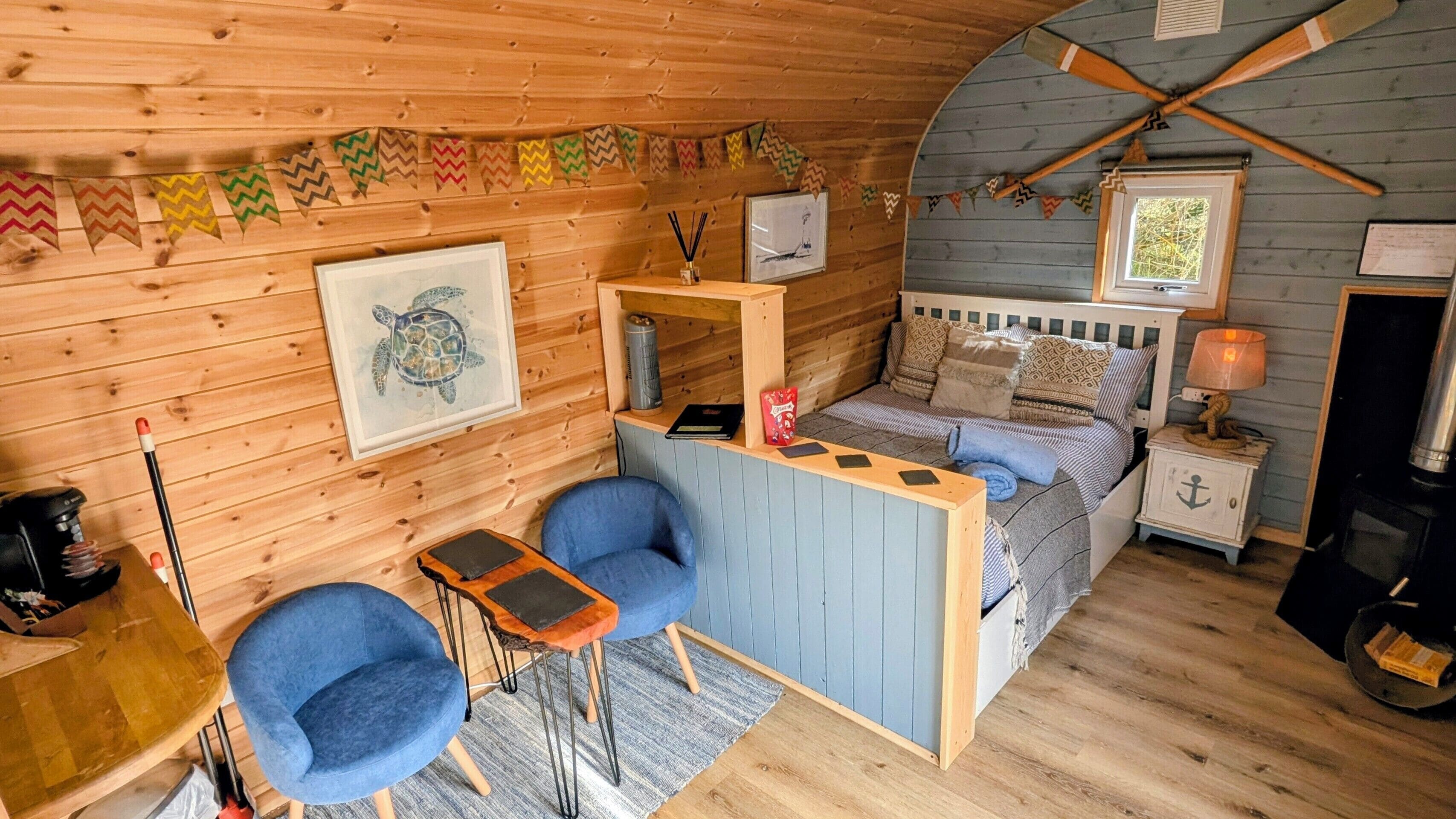 A cozy cabin interior with a wooden interior, a bed with patterned bedding, a desk with a chair, and a small table with a lamp.