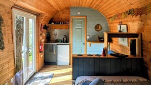 A compact, wooden cabin interior with a kitchenette, a bed, and a door leading to a balcony. - Emlyn's Coppice - Woodland Glamping (Holywell)