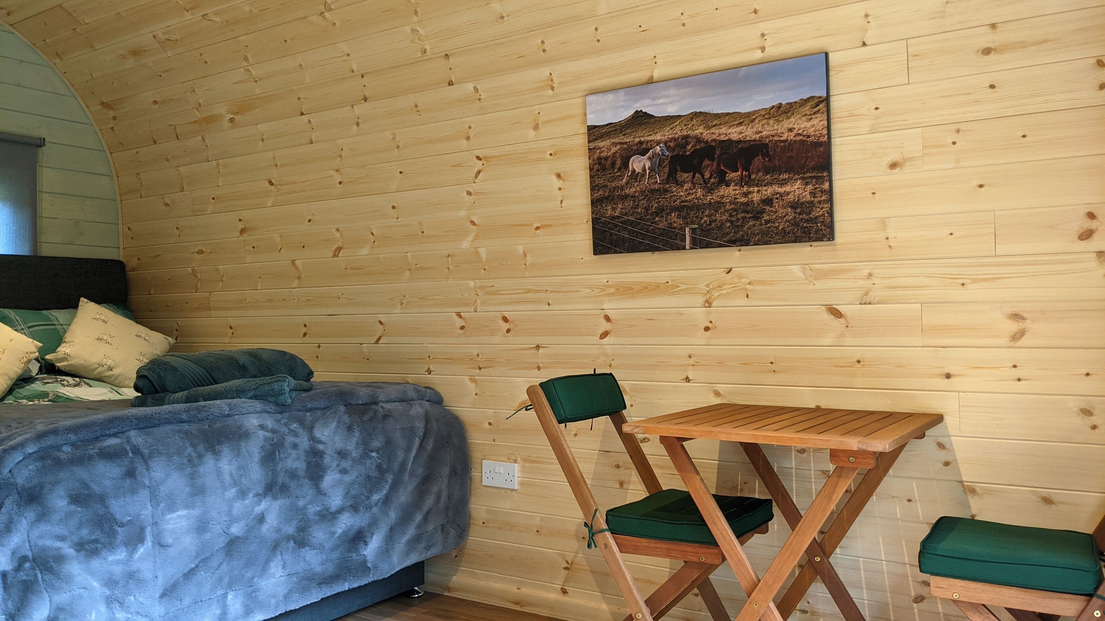 A cozy room with a bed, a small table, and chairs, featuring wooden walls and a mounted picture of horses.