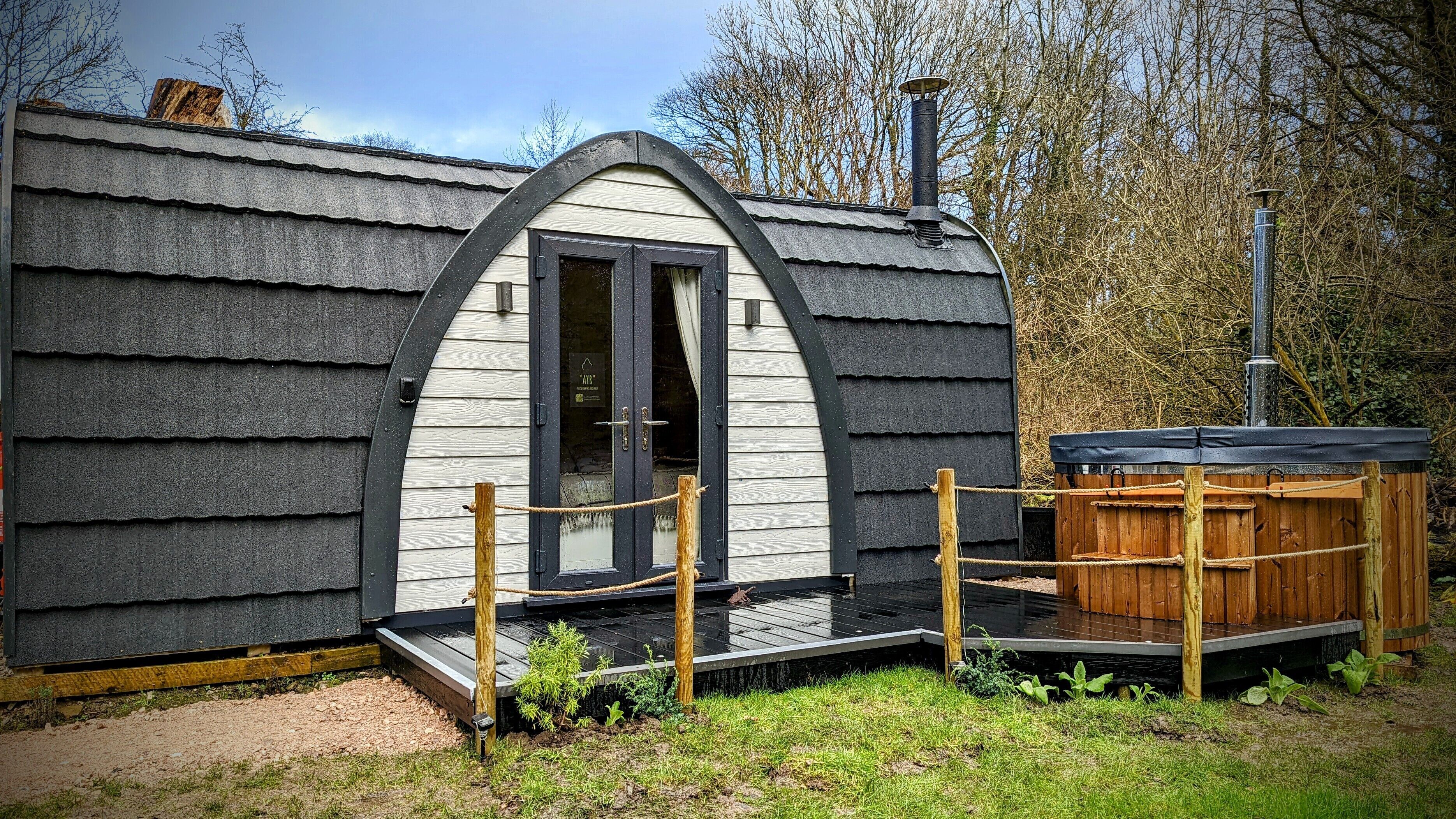 A small, single-story cabin with a sloped roof, a wooden deck, and a hot tub.