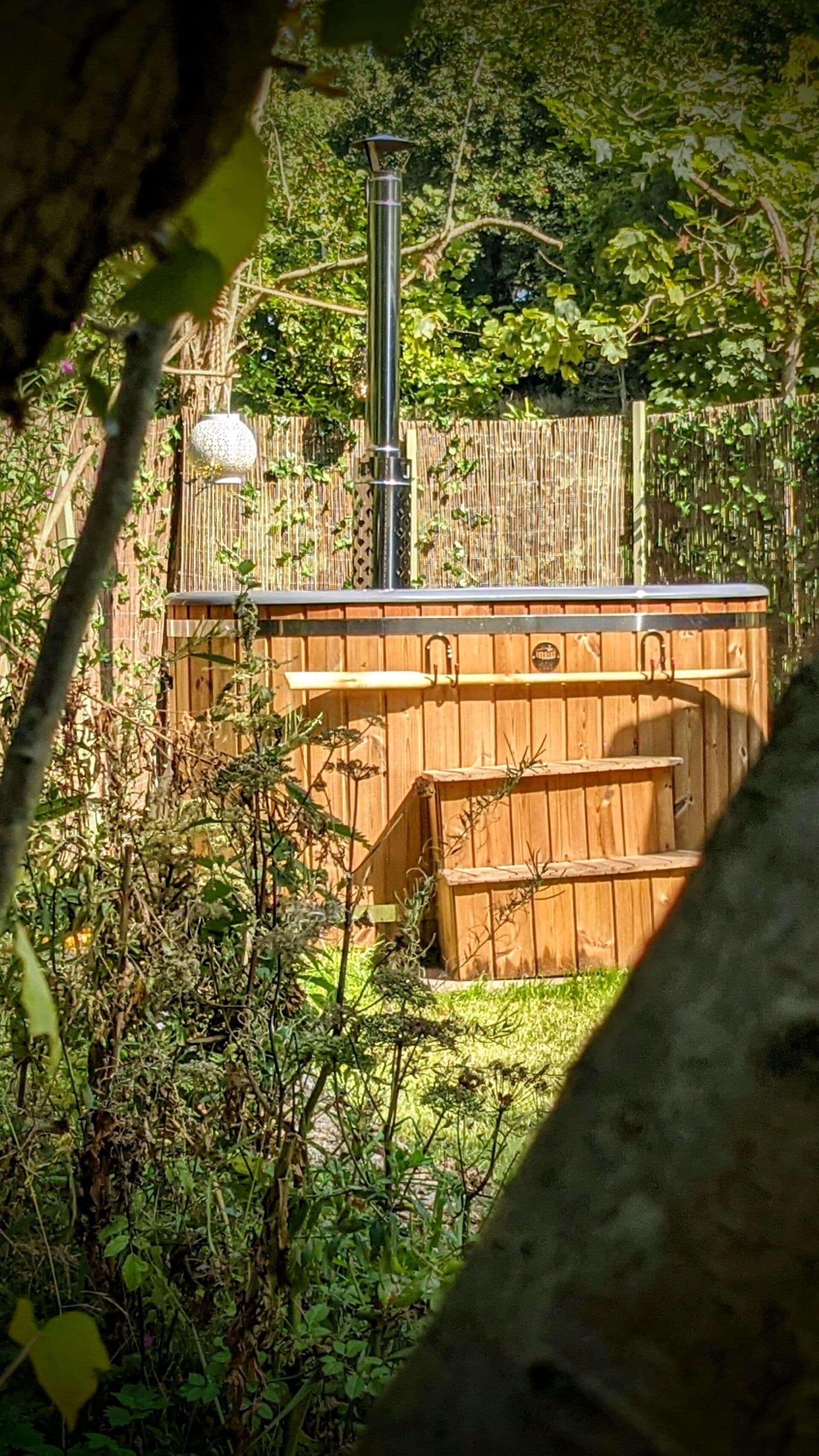A wooden hot tub with a metal frame, surrounded by dense foliage and a bamboo fence.